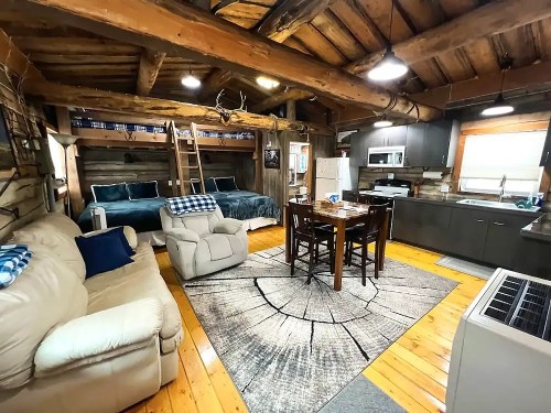 Cozy cabin interior featuring a living area, kitchen, dining table, and lofted sleeping area with wooden beams and rustic decor.