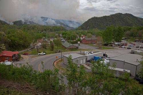 A scenic view of a small town surrounded by mountains, with smoke rising in the distance and greenery in the foreground.