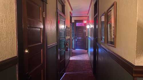 A dimly lit hallway with wooden doors and framed pictures, leading to an exit at the end.