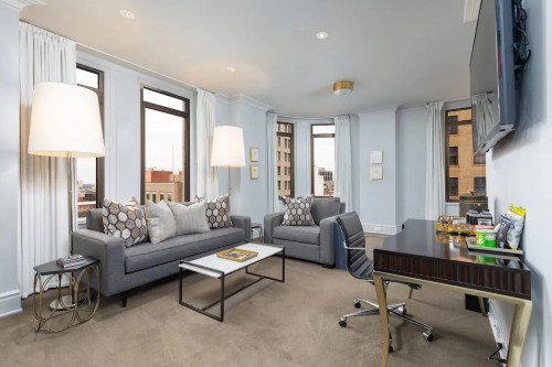 A modern living room with two gray sofas, a coffee table, and a desk, featuring large windows and soft lighting.