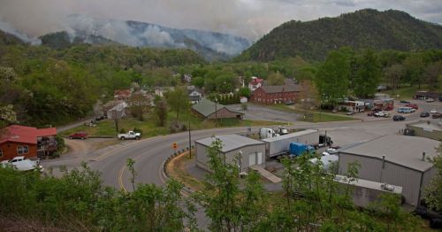 A scenic view of a small town surrounded by mountains, with smoke rising in the distance and greenery throughout.