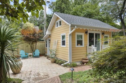 A charming yellow cottage surrounded by greenery, featuring a patio with blue chairs and a brick pathway.