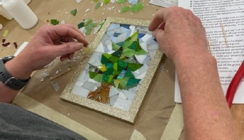 A person assembling a mosaic art piece featuring a tree design, surrounded by colorful glass pieces and tools.