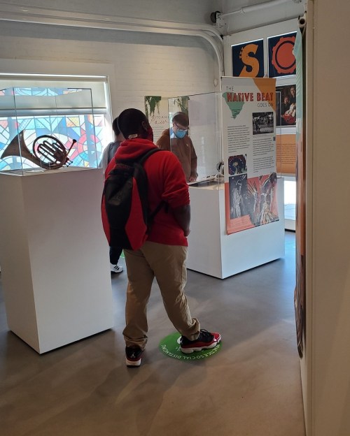 A person in a red hoodie stands on a green marker in a museum, observing an exhibit with informational panels.