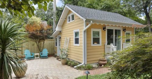 A charming yellow cottage with a porch, surrounded by greenery and a brick patio with blue chairs.