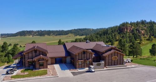 A large wooden lodge with a metal roof, surrounded by green hills and trees under a clear blue sky.