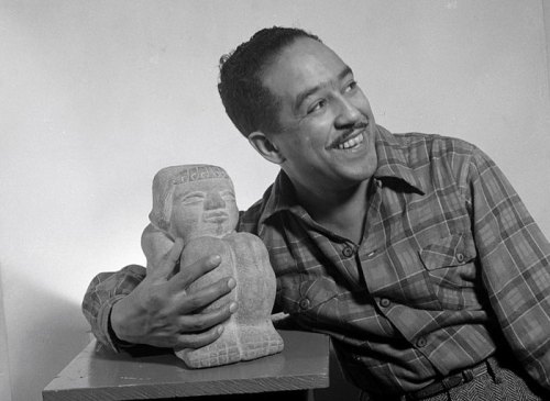 A smiling man in a plaid shirt holds a stone sculpture, posing against a plain background.
