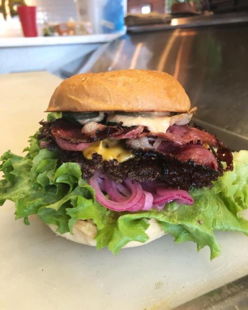 A stacked burger with lettuce, pickled onions, bacon, and sauce on a toasted bun, resting on a countertop.