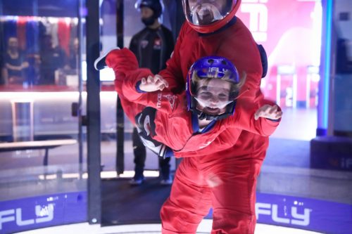 A person in a red jumpsuit is flying in a wind tunnel, being supported by an instructor in a similar outfit.