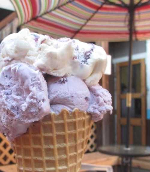 A close-up of a waffle cone filled with three scoops of purple and white ice cream, set against a colorful background.