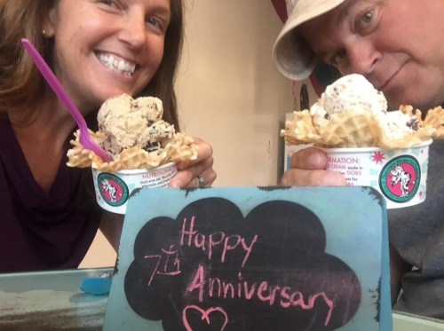 A couple smiles while holding ice cream cones, with a sign that reads "Happy 7th Anniversary" in front of them.