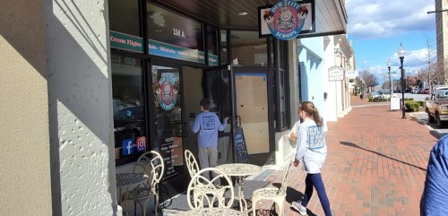 A child and an adult walk towards an ice cream shop with outdoor seating on a sunny day.