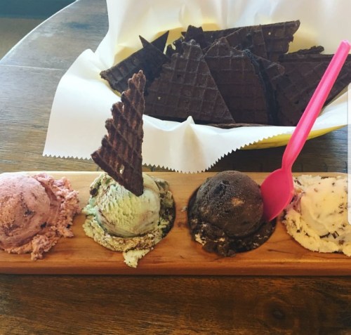 A wooden platter with four scoops of ice cream in pink, green, brown, and white, alongside a bowl of chocolate waffle cones.