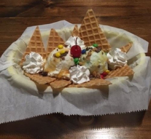 A dessert bowl with two scoops of ice cream, whipped cream, a cherry, and waffle cone pieces.