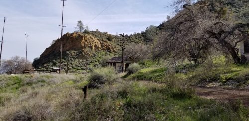 A rustic cabin surrounded by tall grass and rocky hills under a clear sky, with power lines in the background.