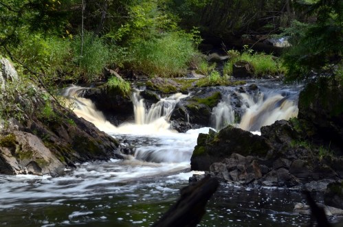 A serene river flows over rocky terrain, surrounded by lush greenery and gentle waterfalls.