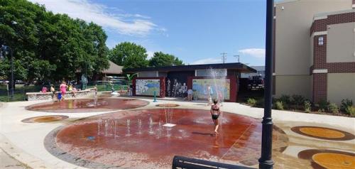 Children play in a splash pad with water features, surrounded by greenery and colorful murals on nearby buildings.