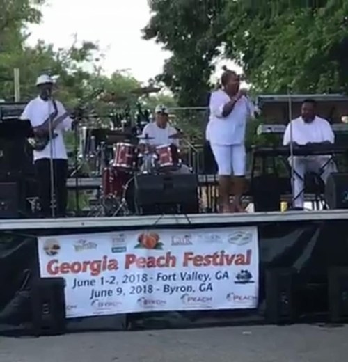 A live band performs on stage at the Georgia Peach Festival, with musicians in white attire and a banner in front.