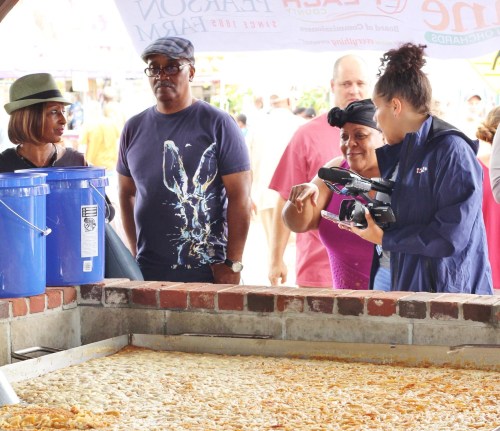 A group of people at a market, observing food preparation, with one person filming and others engaged in conversation.