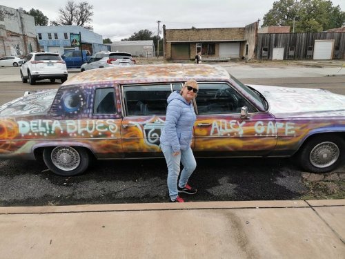 A woman poses next to a colorful, graffiti-covered car with "Delta Blues" and "Alley Cafe" painted on it.