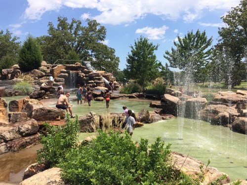 Children and adults play in a natural water feature with rocks and fountains under a sunny sky.