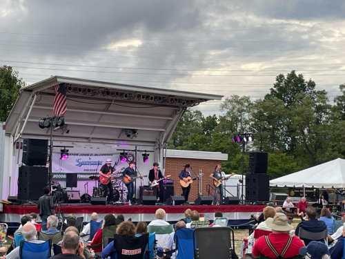 A band performs on stage at an outdoor event, with an audience seated in front on a grassy area.