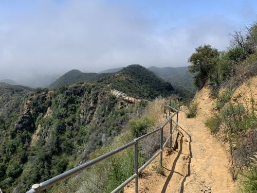 A winding dirt path along a hillside with lush greenery and foggy mountains in the background.