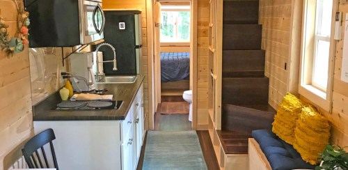 Cozy tiny home interior featuring a kitchen, living area, and stairs leading to a lofted bedroom. Warm wood accents throughout.