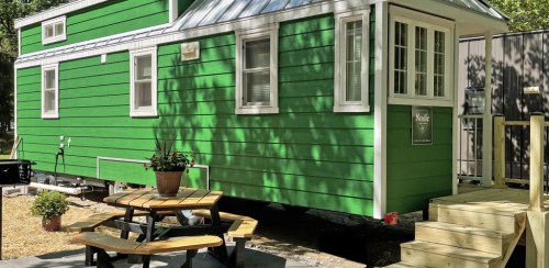 A bright green tiny house with a metal roof, surrounded by greenery and a picnic table in front.