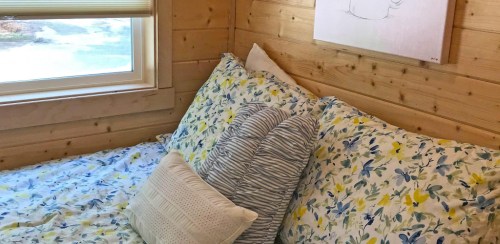 Cozy bedroom with a floral-patterned bedspread, decorative pillows, and a wooden wall. A drawing hangs above the bed.