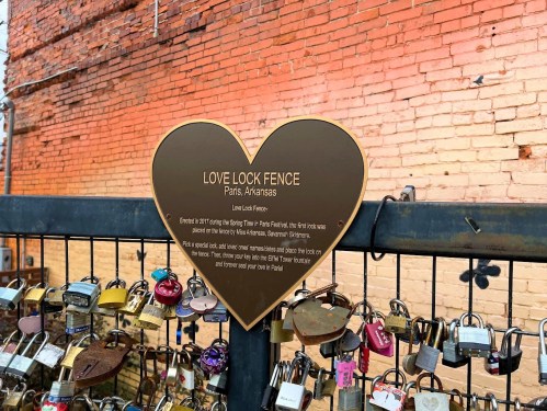 A heart-shaped sign on a fence in Paris, Arkansas, explains the Love Lock Fence tradition with various padlocks attached.