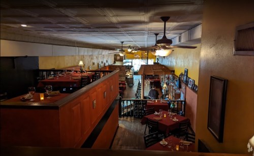 View of a cozy restaurant interior with red tablecloths, wooden accents, and a bar area, featuring warm lighting.