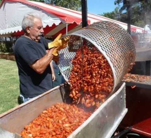 A man in a black shirt pours cooked crawfish from a large basket into a metal container at an outdoor event.