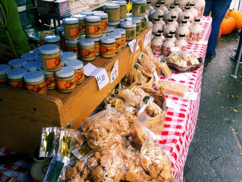 A market stall displaying jars of honey, maple syrup bottles, and various baked goods on a checkered tablecloth.