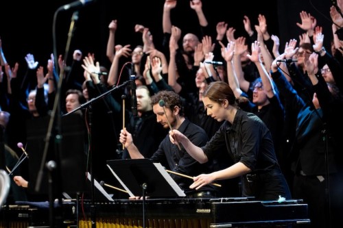 A choir with raised hands performs behind musicians playing a vibraphone and marimba on stage.