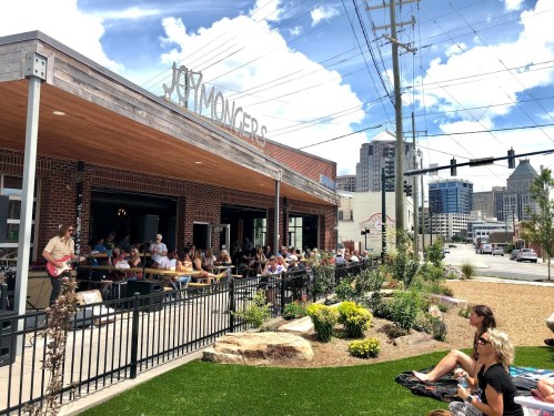 A bustling restaurant patio filled with diners, surrounded by greenery and city buildings under a bright blue sky.