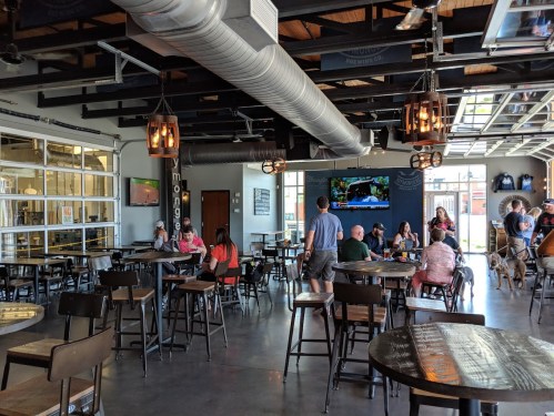 A lively bar interior with wooden tables, patrons enjoying drinks, and a TV showing a sports game.