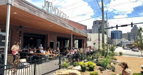 A bustling restaurant patio filled with people enjoying their meals, with a city skyline in the background under a blue sky.