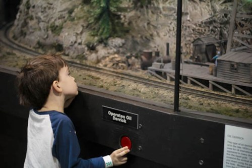 A child curiously observes a miniature oil derrick display at a museum, with a button labeled "Operates Oil Derrick."