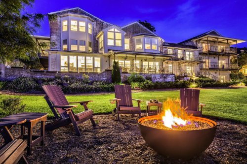 A cozy outdoor scene featuring a fire pit surrounded by chairs, with a large house illuminated at dusk in the background.