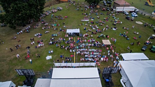 Aerial view of a large crowd gathered on a grassy field for an outdoor event, with tents and seating areas visible.