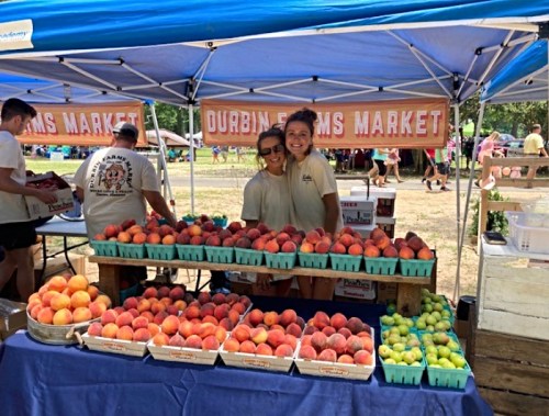 Two smiling vendors stand behind a table filled with fresh peaches and other fruits at a market stall.