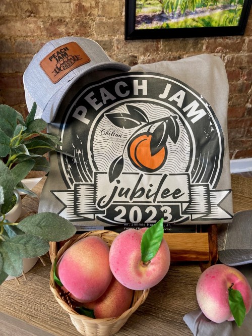 A gray cap and a t-shirt with a "Peach Jam Jubilee 2023" design, surrounded by fresh peaches and greenery.