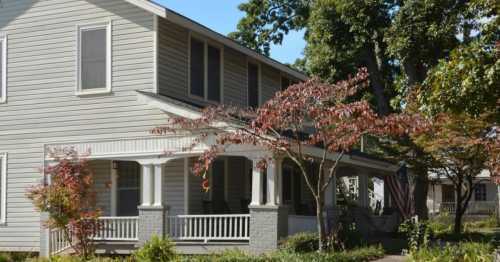 A two-story house with a porch, surrounded by trees and colorful shrubs on a sunny day.