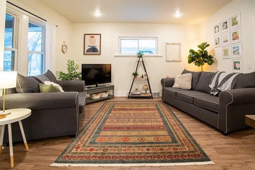 Cozy living room with gray sofas, a TV, a decorative rug, and wall art, featuring plants and warm lighting.