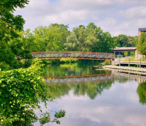 A wooden bridge spans a calm river, surrounded by lush greenery and reflecting clouds in the water.