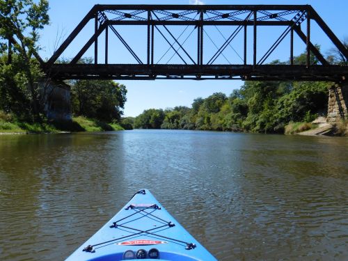 A kayak view of a calm river beneath an old metal bridge, surrounded by lush greenery and clear blue skies.