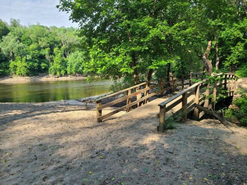 A wooden bridge over a calm river, surrounded by lush green trees and sandy banks under a clear blue sky.