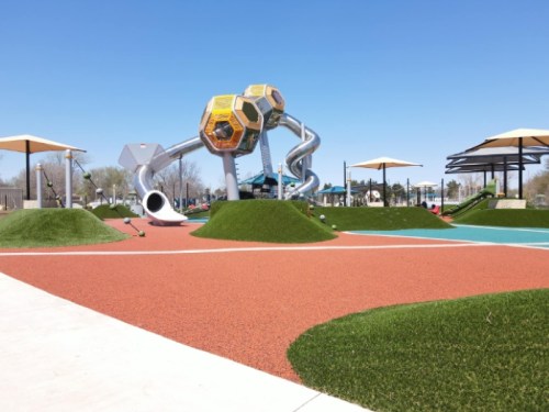 Colorful playground with a large slide, climbing structures, and green hills under a clear blue sky.