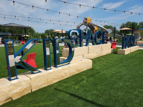 Colorful "KID SPACE PARK" sign on a grassy area, with playground equipment and string lights in the background.